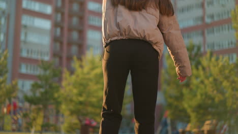 a back shot of a girl wearing a peach jacket and black trousers rollerblading through a park.surrounded by greenery and park benches, with tall urban buildings in the background