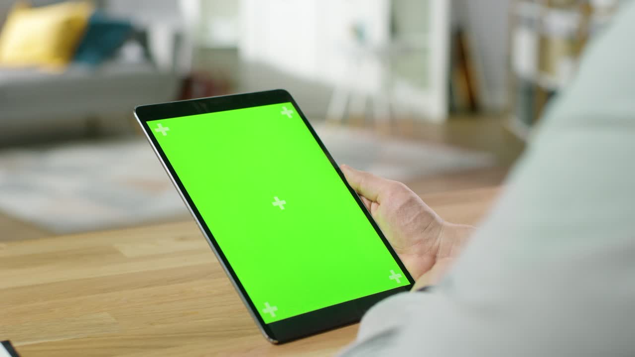 Mand bruger grøn mock-up skærm tablet i portrættilstand, mens han sidder ved sit skrivebord. I baggrunden hyggelig stue