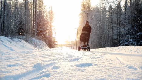 Toma-En-Cámara-Lenta-De-ángulo-Bajo-De-Un-Hombre-Caminando-Con-Un-Vagón-De-Bebé-En-Un-Clima-Invernal-Nevado,-Con-Un-Hermoso-Sol-Dorado-En-El-Fondo