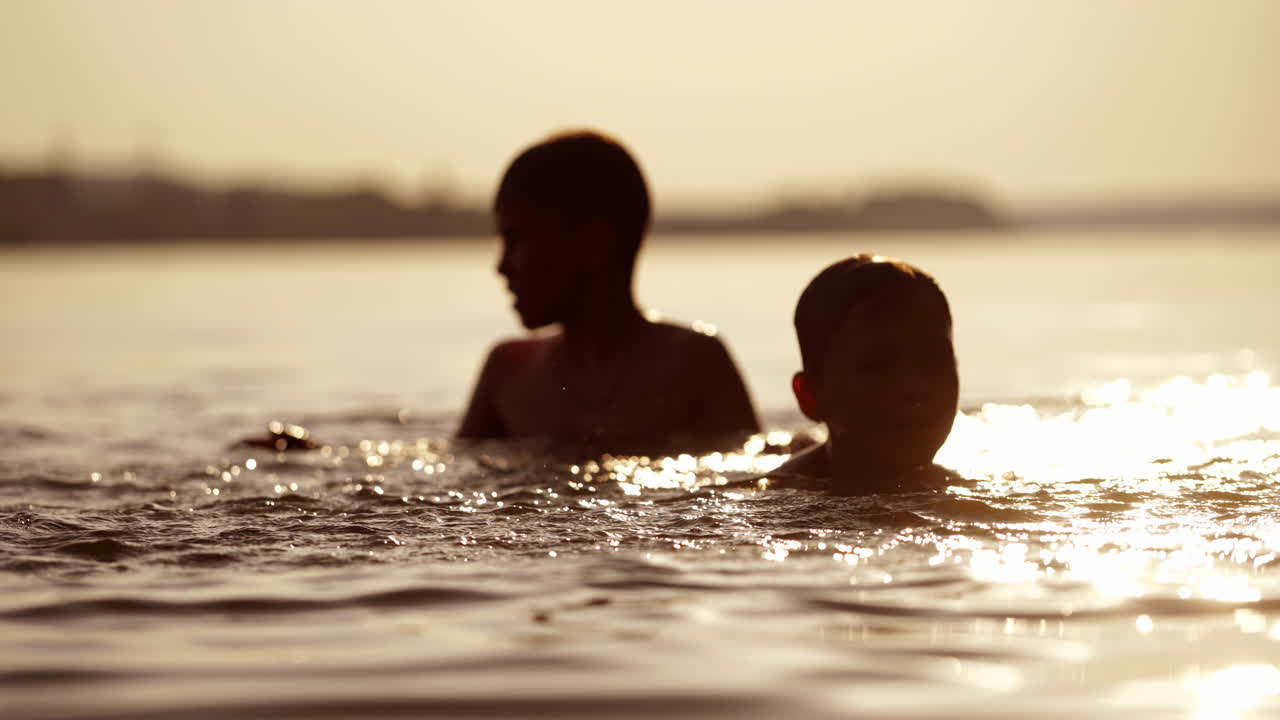 Silhouette of two boys swimming and playing with water at sunset. Happy brothers having good time together in the river in the evening. Close-up.