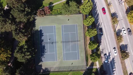 1-2-Luftaufnahme-Von-Oben-Aus-Der-Vogelperspektive-Von-Paaren,-Die-Im-Heißen-Sommer-Tennis-Spielen,-In-Einem-Modernen-Park-In-Der-Nähe-Eines-Luxuriösen-Viertels-In-Der-Nähe-Einer-Gartenstraßenautobahn-Mit-Leichtem-Verkehr-In-Der-Tropischen-Heimatstadt