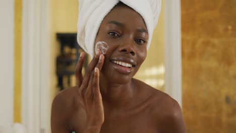 Portrait-of-smiling-african-american-woman-with-towel-using-cream-on-her-face-in-bathroom