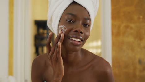 retrato de una mujer afroamericana sonriente con una toalla usando crema en su cara en el baño