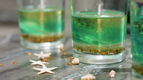 housewife carefully drops small multicolored shell-like and star-shaped decorations onto kitchen countertop beside glass filled with layered green and gold candle mixture