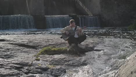 young man playing guitar sitting on the bank of a mountain river on a background of rocks. concept of freedom relaxation. place