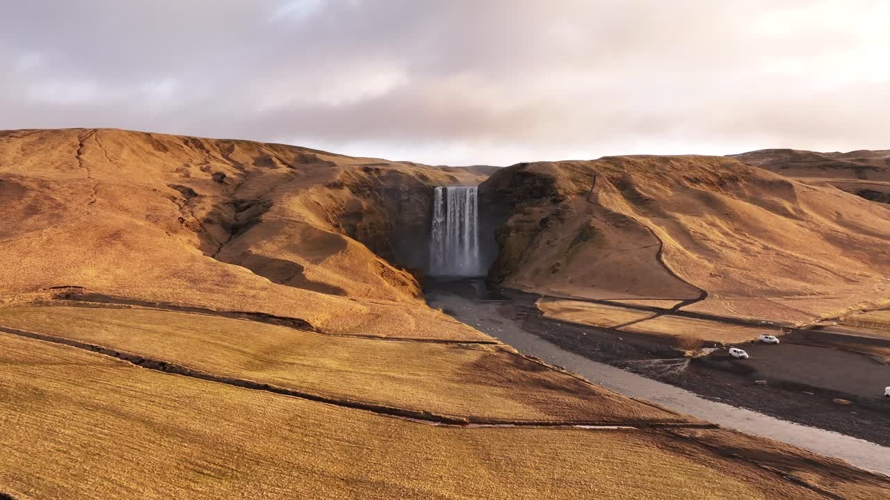 Drone pulls back to reveal Skógafoss waterfall in Iceland, set in a golden valley below Eyjafjallajökull.