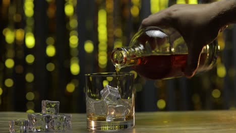 pouring of golden whiskey, cognac or brandy from bottle into glass with ice cubes. shiny background