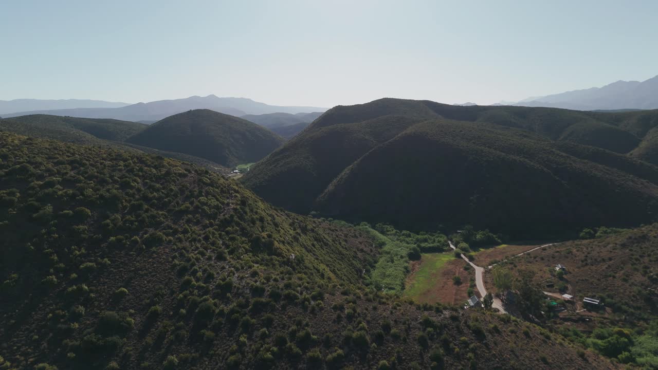 Aerial shot of the Klein Karoo mountain range, Western Cape, South Africa, covered in vibrant fynbos plants on a sunny day. The drone reveals small houses and paddocks nestled in the valley below.