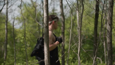 soldier wearing headband and gloves moves through dense forest trunks pushing aside thin branches while carrying backpack strap across shoulder filtering light through foliage onto skin