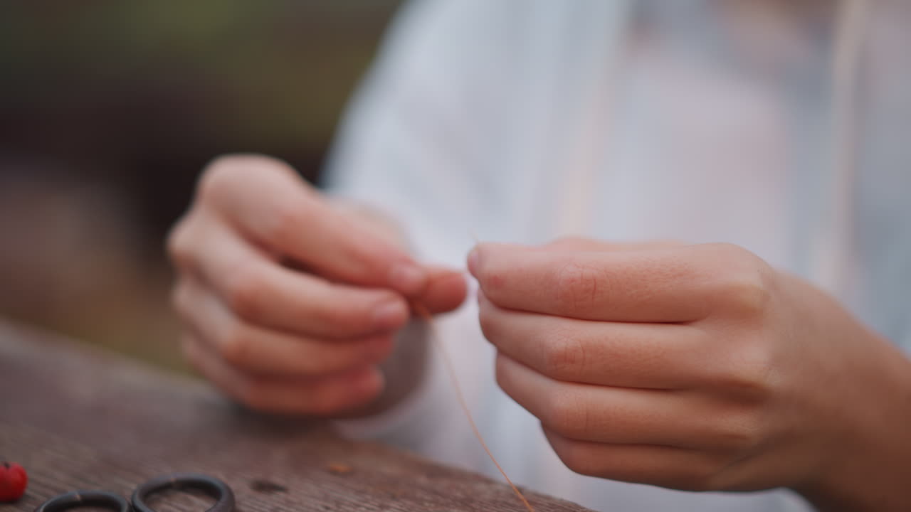 Closeup caucasian hands threading string. Quiet outdoor bench scene with wooden table, small red bead, scissors at edge, slow focused fingers knotting delicate thread, warm glow, tactile texture