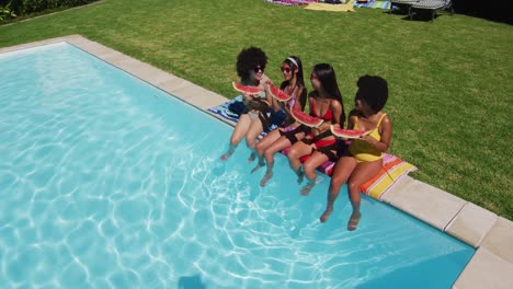 Vista-Aérea-De-Un-Grupo-De-Chicas-Diversas-Comiendo-Sandía-Mientras-Están-Sentadas-Junto-A-La-Piscina