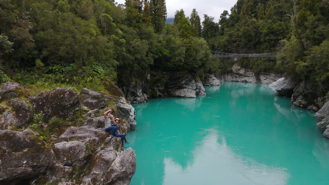 Couple admires the stunning turquoise waters of Hokitika Gorge, surrounded by lush greenery on New Zealand’s West Coast, aerial orbit as they sit on cliff edge
