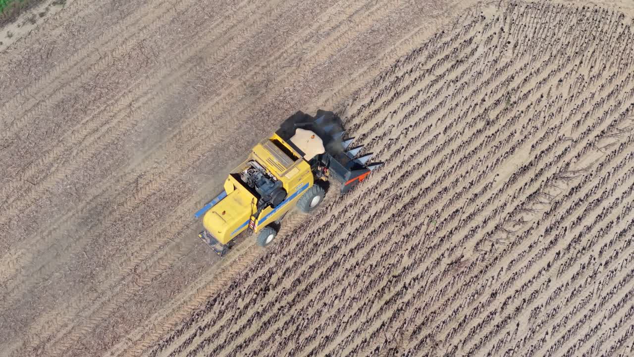 A yellow combine harvester operates in a harvested field, leaving behind rows of cut stalks and dry soil. The aerial view captures the machinery at work during the agricultural harvesting process