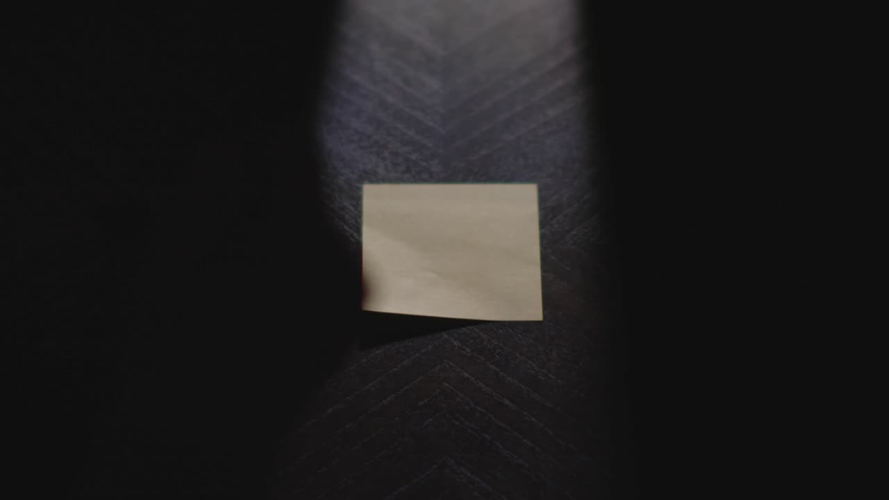 A close up of a hand writing "Monday" on a sticky note with a marker, picking it up, and placing it on a desk in harsh light