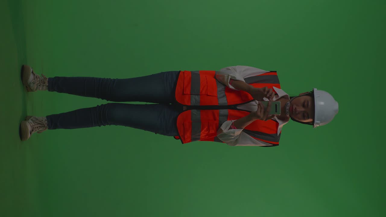 Full Body Of Asian Female Engineer With Safety Helmet Using Smartphone While Standing In The Green Screen Background Studio, During Sunset Or Sunrise Time