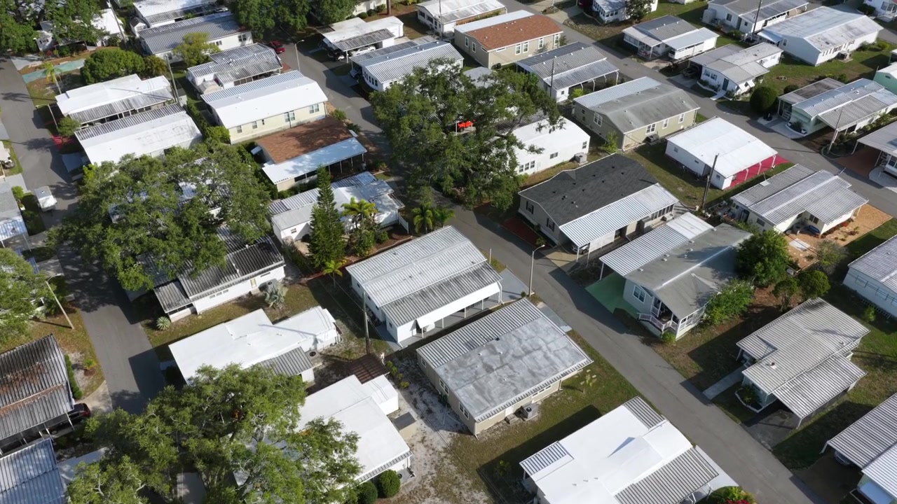 Una fitta griglia di tetti di case mobili riempie il quartiere di Clearwater, querce mature ombreggiano strade strette che si snodano tra case prefabbricate ravvicinate. Ripresa aerea con drone.
