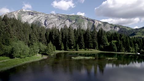 Aerial-of-French-Alps-pine-tree-forest-with-clouds-in-blue-sky-above-mountain-rocks-mirrored-in-Lac-du-Mines-d'Or-[translation:-Lake-of-Gold-Mines]-Tour-de-France-vacation-outdoor-sports-during-summer