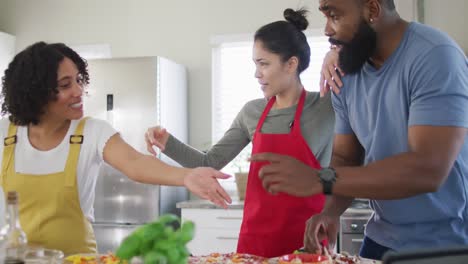 smiling diverse female and male friends making pizza and cooking in kitchen, in slow motion