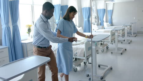 diverse male doctor helping female patient walk with drip strand in hospital ward, slow motion