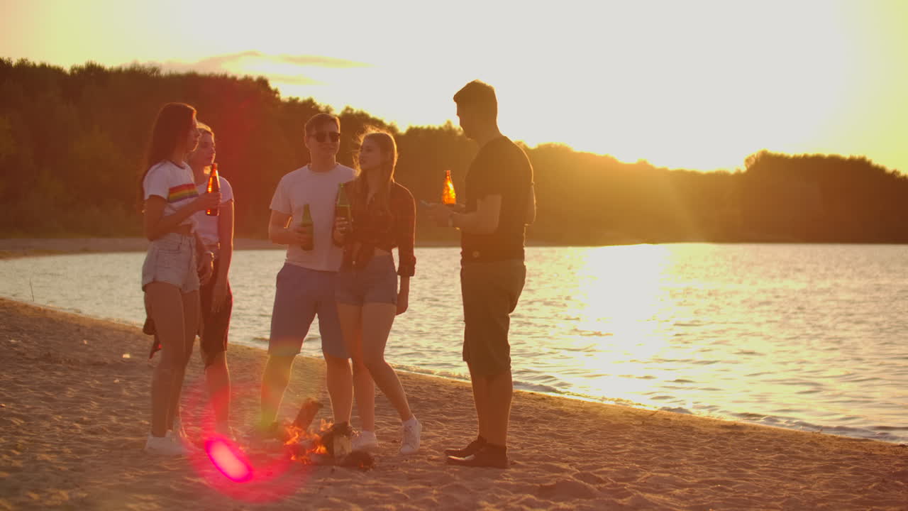 los cinco mejores amigos pasan el tiempo al atardecer en la playa de arena en pantalones cortos y camisetas alrededor de una hoguera con cerveza. están hablando entre sí y disfrutando de la noche de verano en la naturaleza.