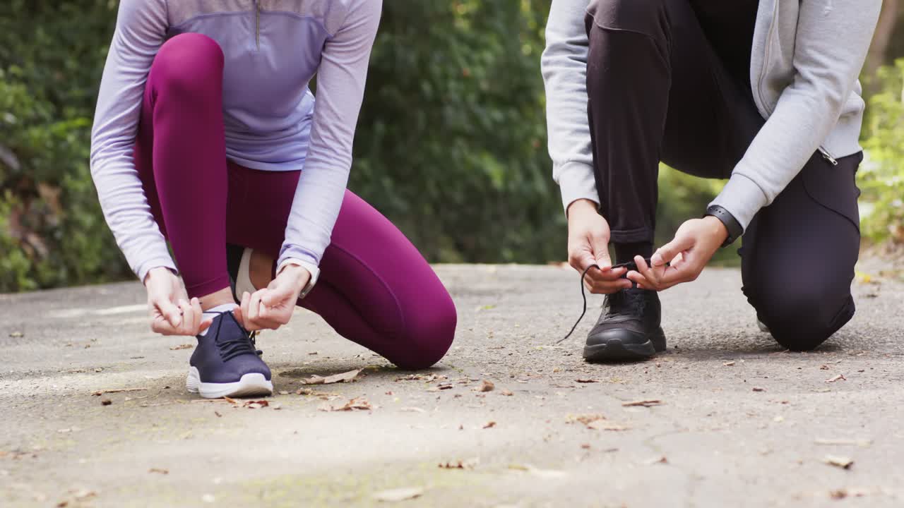 Una coppia in abbigliamento sportivo si inginocchia per allacciarsi le scarpe su un sentiero nel bosco prima di correre.