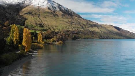 flying over beautiful blue lake wakatipu, queenstown, new zealand with mountains fresh snow, clouds, blue sky autumn trees in the background - aerial drone