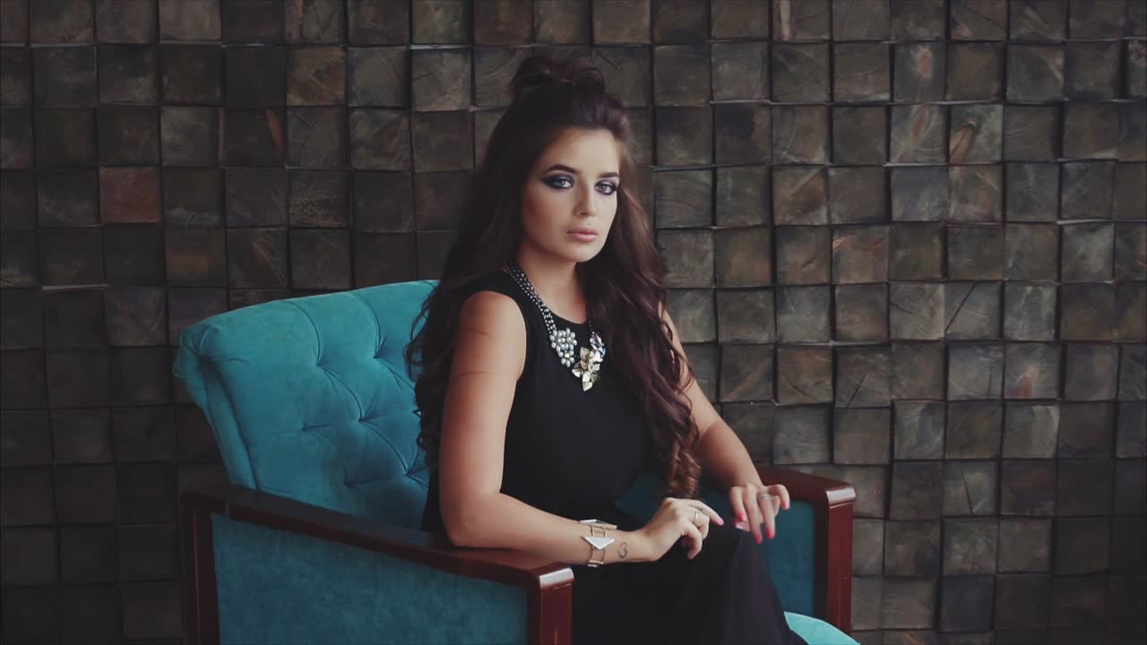 mujer en silla azul, traje negro y joyas