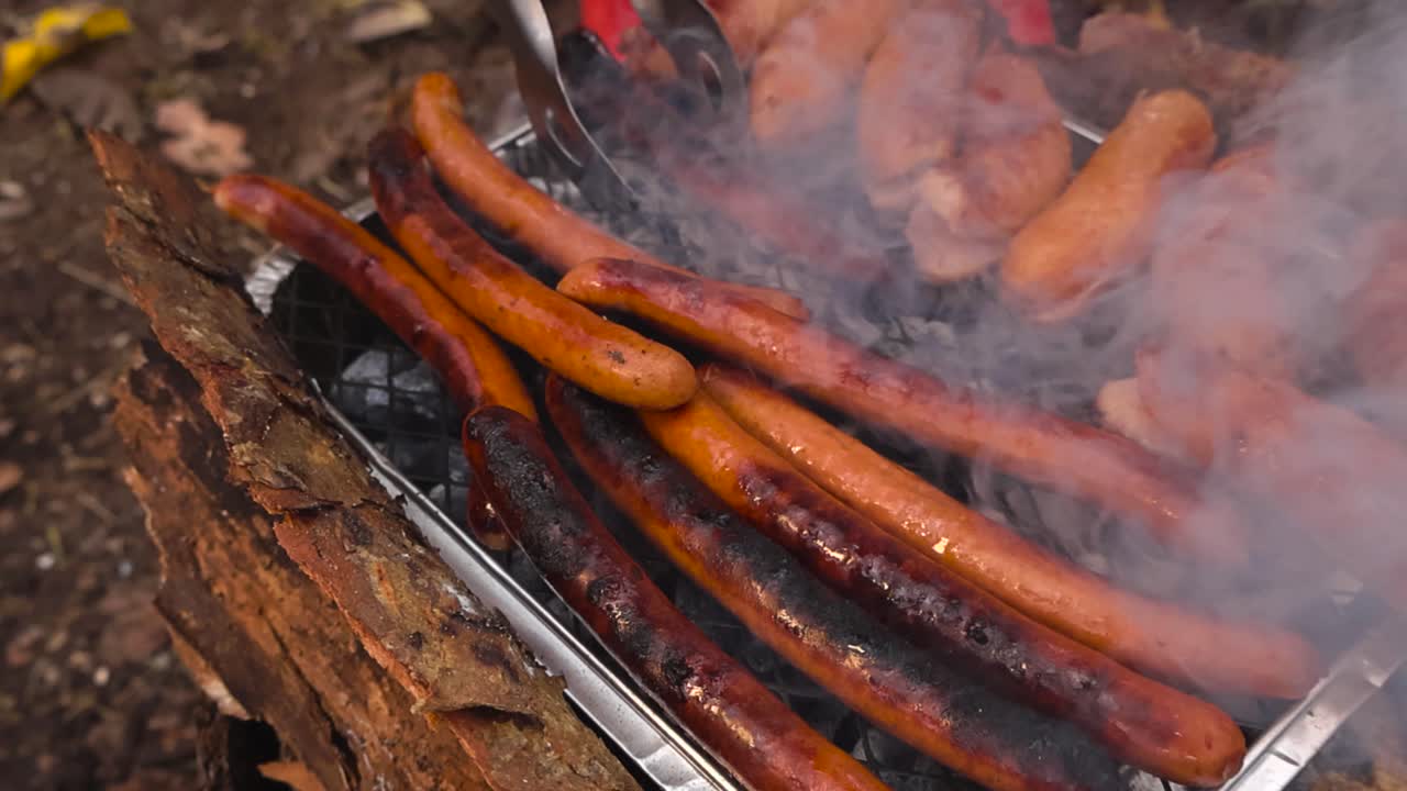 Person with tongs moving golden brown sausages and hot dogs on a instant grill or barbecue that is smoking with hot embers in a autumn garden. White smoke moves in slow motion cinematically