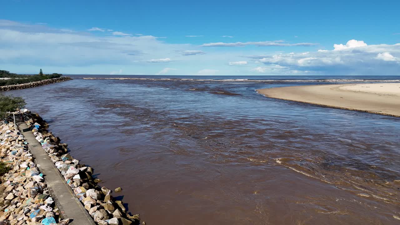Aerial drone footage of brown floodwaters meeting ocean at river mouth, rocky jetty, Australia
