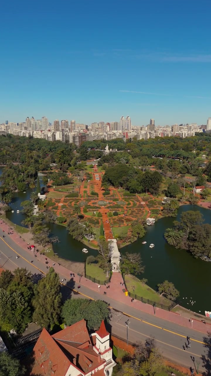 Veduta aerea che si eleva sopra un parco cittadino con giardini, laghi e un campo sportivo in una giornata di sole.