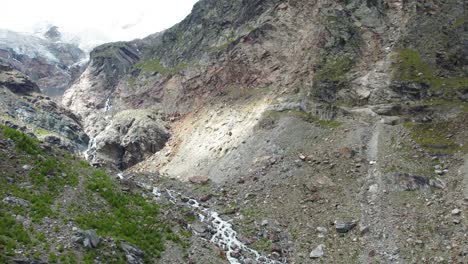 Impresionante-Toma-Aérea-De-Una-Formación-Montañosa-Masiva-En-Saas-Fee-Con-Un-Río-Salvaje-Con-Cascada-Y-Un-Glaciar,-Alpes-Suizos