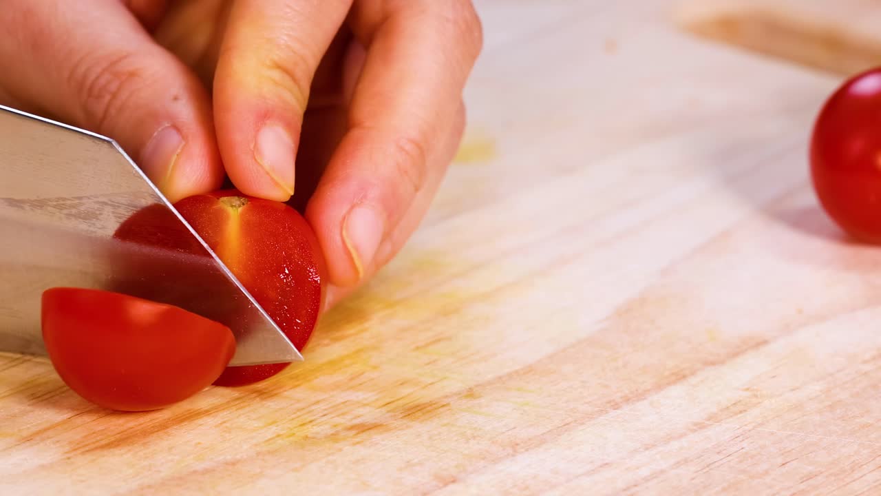 A hand precisely slices cherry tomatoes on a wooden cutting board using a sharp knife.