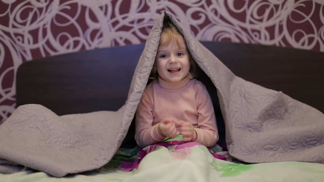 Piccola bambina allegra si nasconde sotto la coperta e guarda la TV. Concetto di sonno infantile.