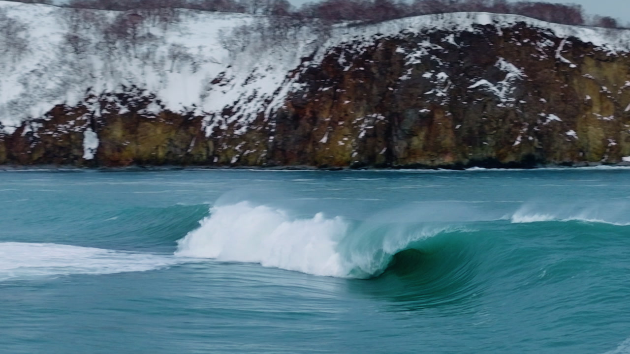 Clean turquoise wave breaking near rugged snow covered coastal cliffs in cold ocean water, empty fast breaking powerful wave