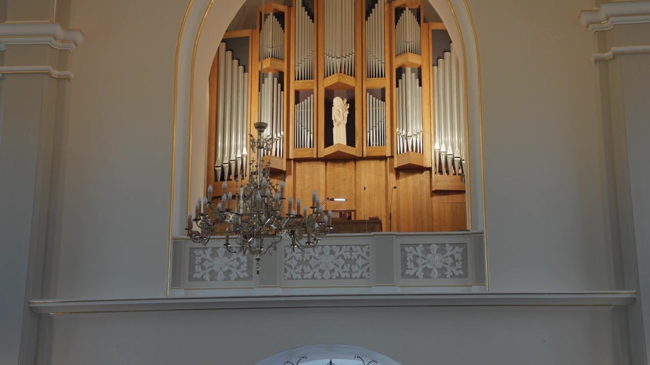 Wide angle shot showcases a grand pipe organ and glowing chandeliers in a serene, softly lit church. Intricate architectural details highlight the calm, reverent atmosphere. Nobody visible.