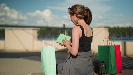 back view of a woman seated on an outdoor bench with shopping bags on both sides, she retrieves a book from a mint green bag, opens it, and flips through its pages