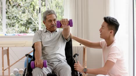 diverse male physiotherapist advising and senior man in wheelchair using dumbbells, in slow motion