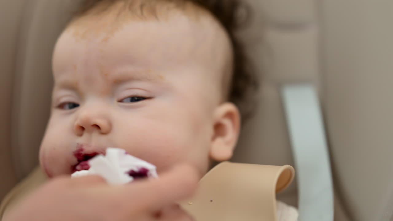 Infant seated in a high chair while an adult feeds the baby with a spoon