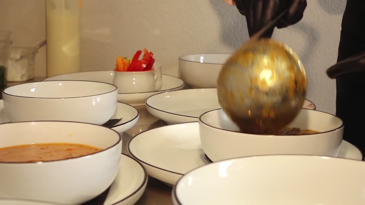 Várias tigelas de sopa quente servidas com pão torrado em pratos em uma cozinha de restaurante, com batatas fritas, salada e vegetais em conserva visíveis, enquanto uma mão enluvada serve os pratos.
