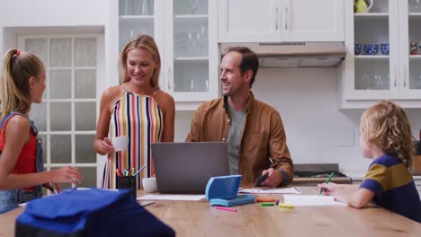 caucasian parents embracing in kitchen, father working on laptop and children doing school work