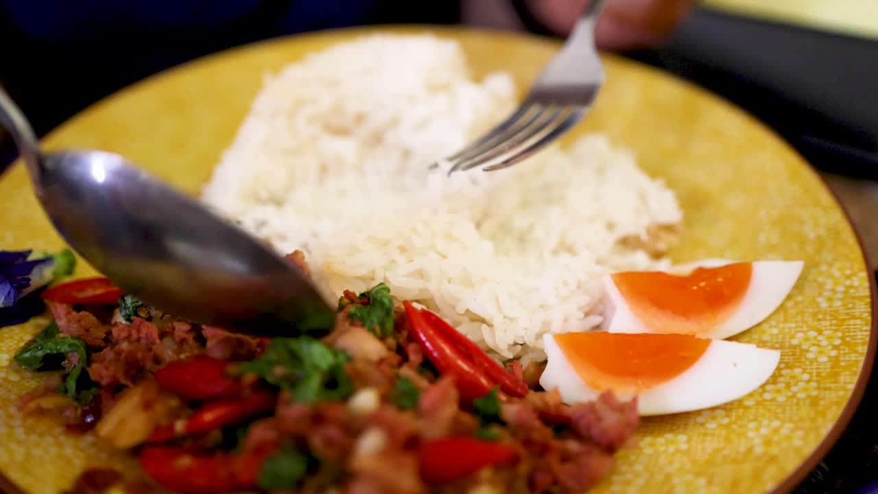 A close-up sequence shows a person using a spoon and fork to mix steamed rice, boiled egg, and fermented pork sausage on a yellow plate under warm lighting