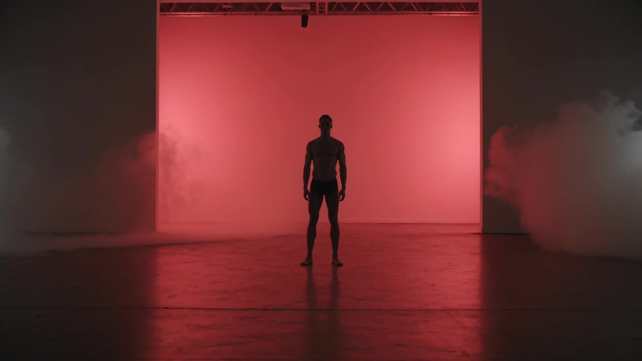 Homem descalço de shorts posando diante de um fundo vermelho, máquinas de fumaça liberando névoa para drama
