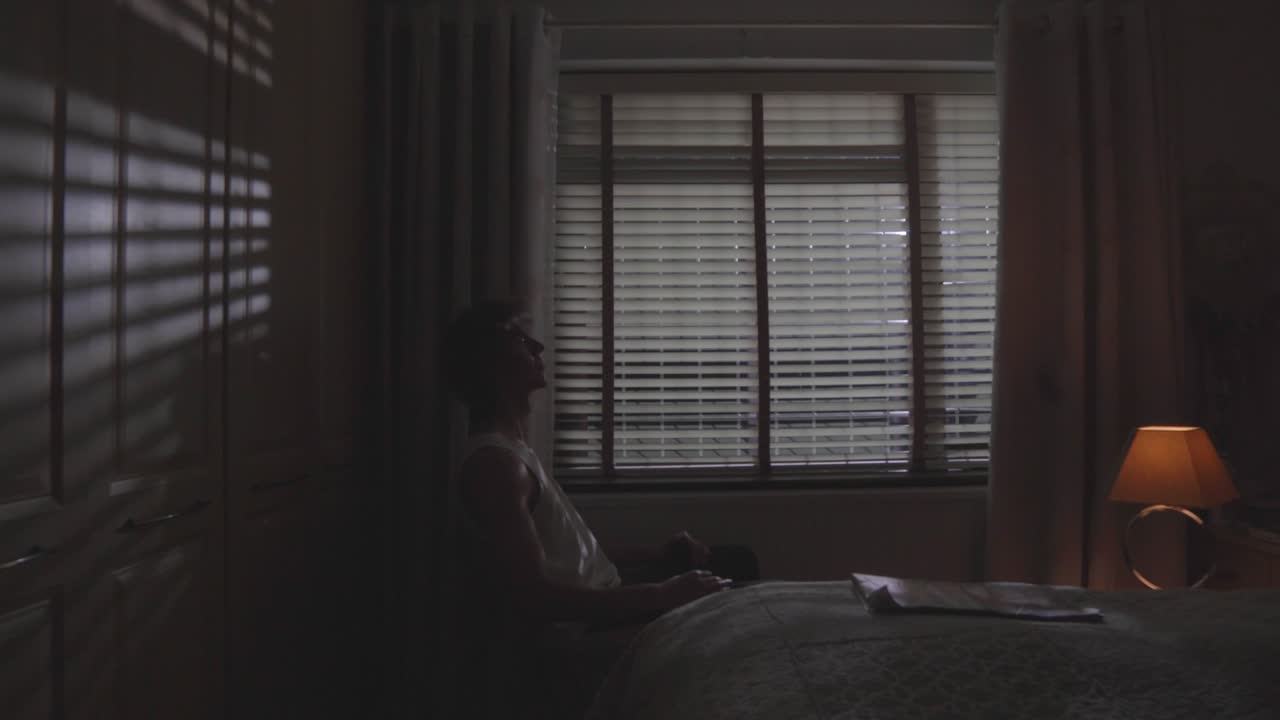 Over-the-shoulder view of man sitting alone at desk in moody room lit by window light, side angle static as he leans against wall