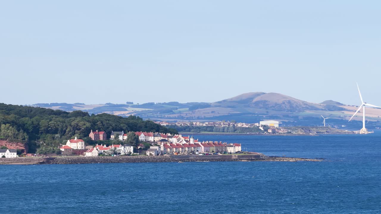 Wide shot of seaside village, wind turbine, and hills under clear daylight in Fife, Scotland