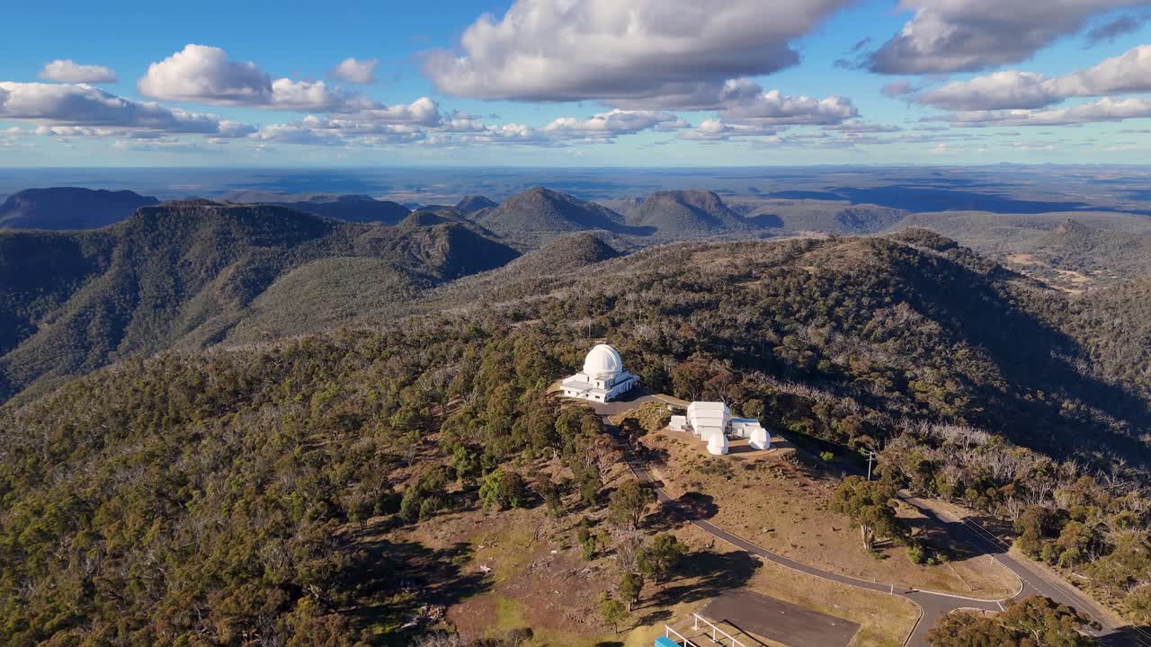 Aerial footage captures a white observatory dome atop forested mountains under dramatic clouds, with smooth camera movement revealing expansive natural landscapes and scientific structures