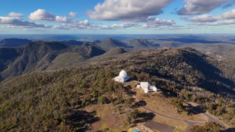 aerial footage captures a white observatory dome atop forested mountains under dramatic clouds, with smooth camera movement revealing expansive natural landscapes and scientific structures