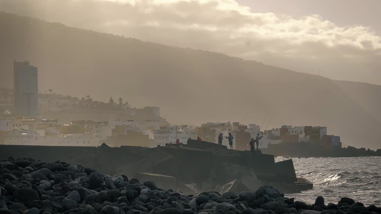 People fishing off the rocky shore of Tenerife, Spain on a peaceful hazy morning with volumetric rays of light beaming through the clouds as the splashing waves emerge from the rocks in slow motion.