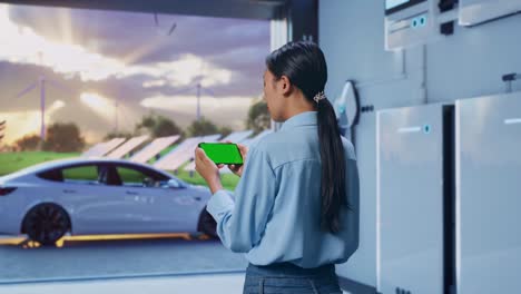 back view of asian female with home energy storage system in a modern garage, checking on smartphone with green screen and look at the factory