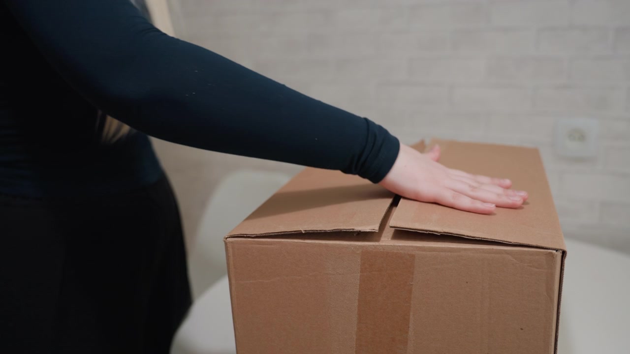 Caucasian woman closing and lifting box, hands flattening flaps then sealing cardboard, carrying load across empty dining area with round table and white chair, light brick wall backdrop, practical