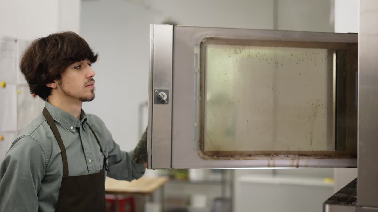 Young man baker in apron and glove taking fresh pastry out of oven in bakery kitchen. Side view of male chef opening oven and taking tray with tasty pies cooking in modern kitchen
